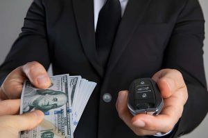 Cash for cars near me—man in suit handing car key fob while receiving stack of hundred-dollar bills.