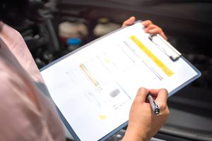 Car sales Sarasota FL dealer completing vehicle inspection checklist with clipboard near engine bay.